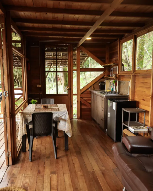 Comedor y cocina de la cabaña, con piso, techo y paredes de madera y ventanas hacia el bosque.
